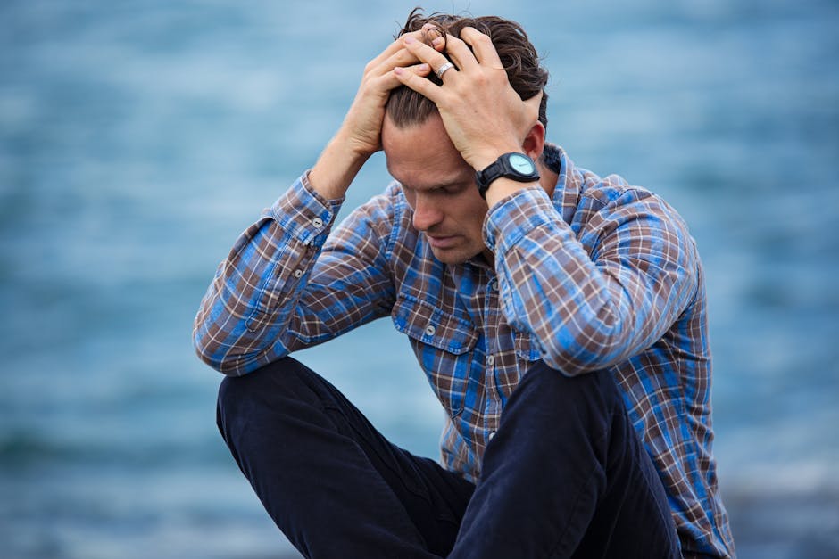 A man in a plaid shirt sits by the water, reflecting on mental health support coaching and emotional well-being